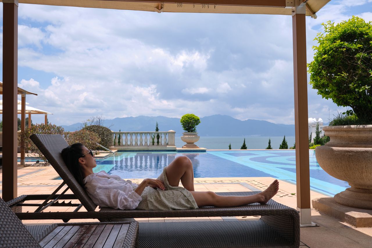 gallery-1 A woman in sunglasses relaxes on a sunlounger by a luxury poolside with a scenic view.