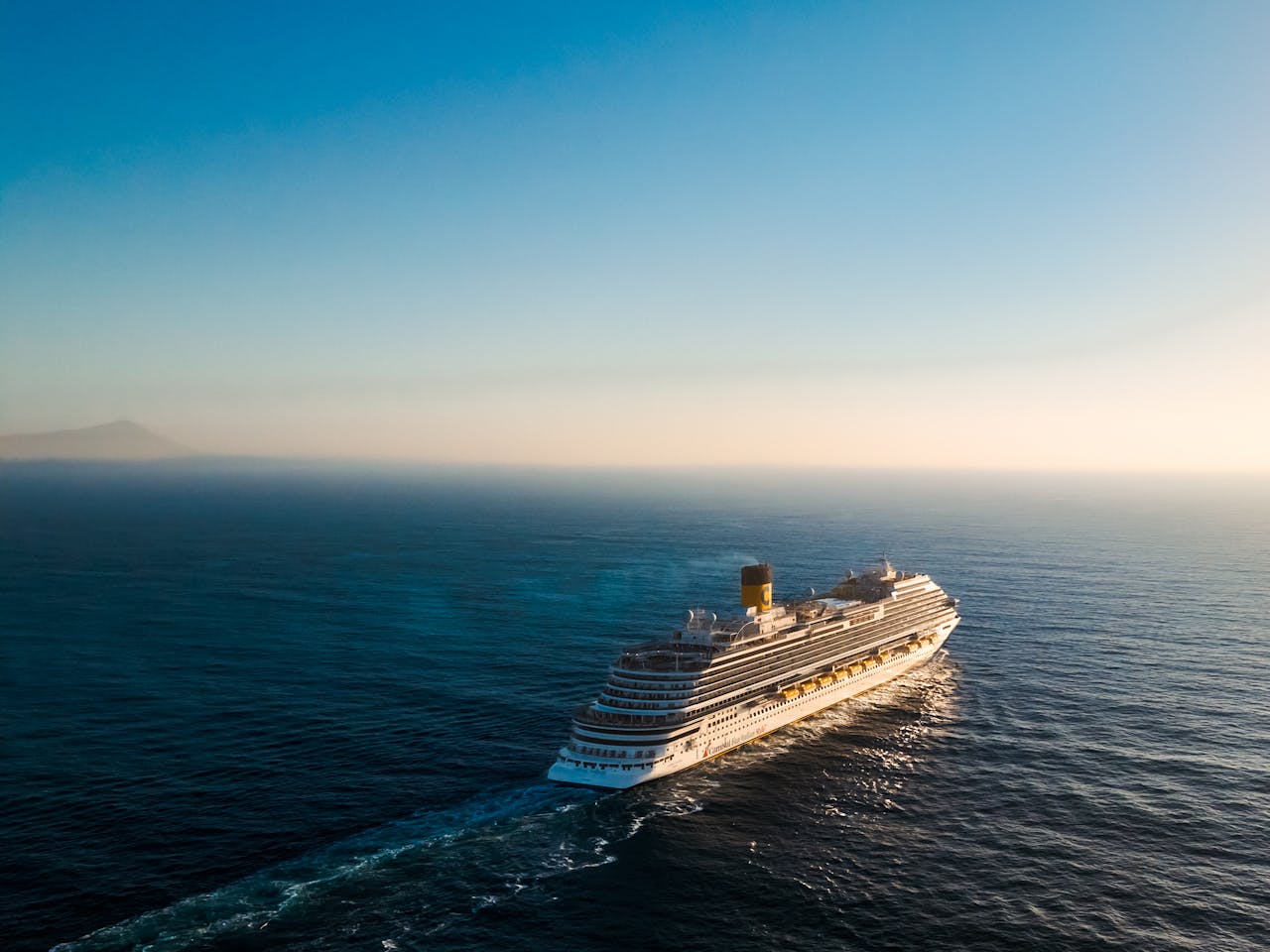 hero-img-01 Aerial view of a luxury cruise ship navigating the calm ocean waters during sunset.